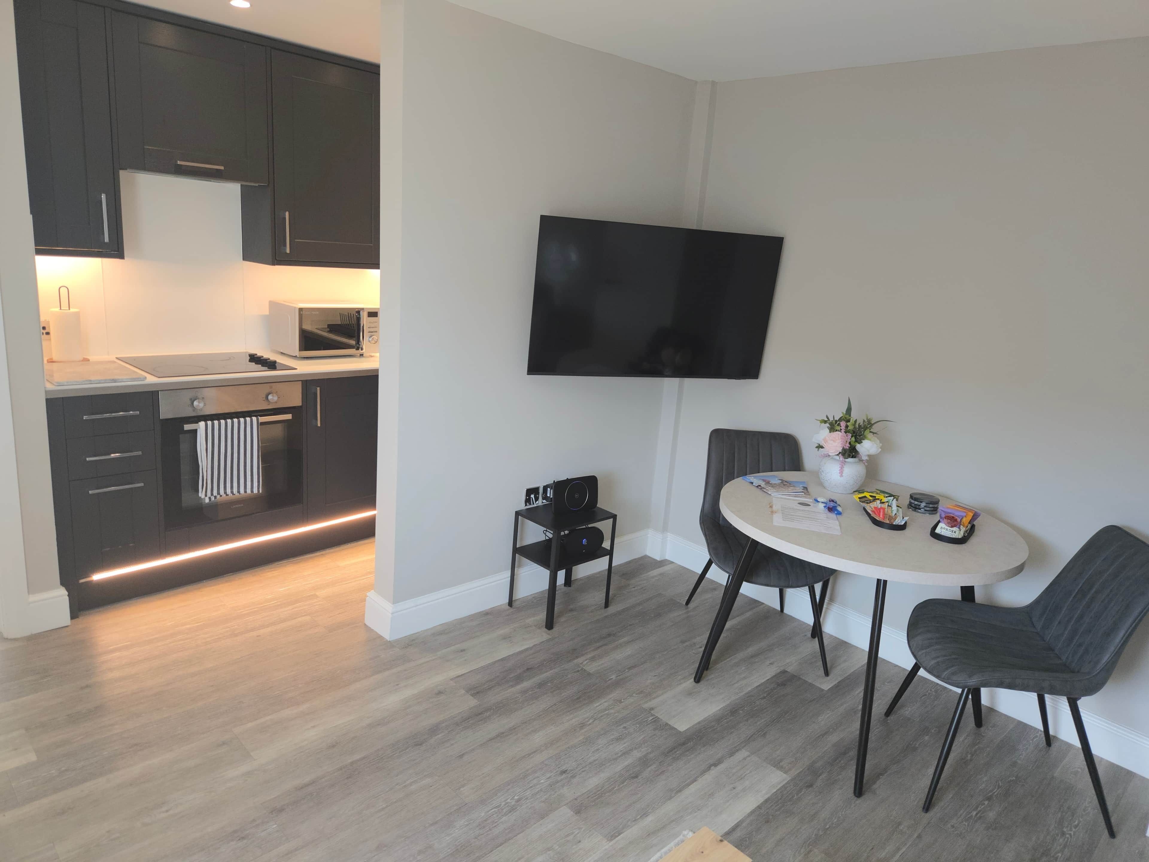 Una cocina moderna con armarios oscuros, un microondas y un horno integrado. Hay una zona de comedor con una mesa redonda, dos sillas y un televisor de pantalla plana montado en la pared.