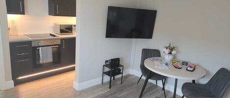 A modern kitchen with dark cabinetry, a microwave, and a built-in oven. There is a dining area with a round table, two chairs, and a flat-screen TV mounted on the wall.
