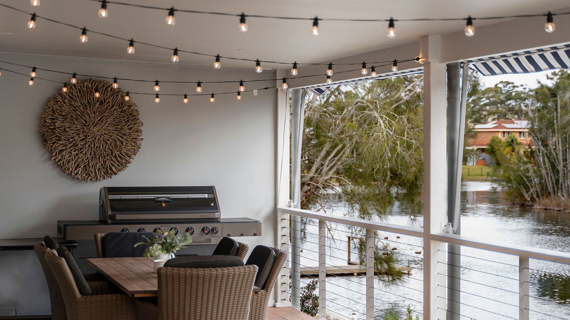 An outdoor dining area with a grill, wicker furniture, string lights, and a view of a water body with trees.