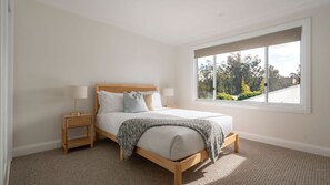 A bedroom with a large window, a bed with white bedding, two bedside tables with lamps, and a view of trees outside. - Holiday Mode | Lakefront Luxury with private jetty (Lake Conjola)