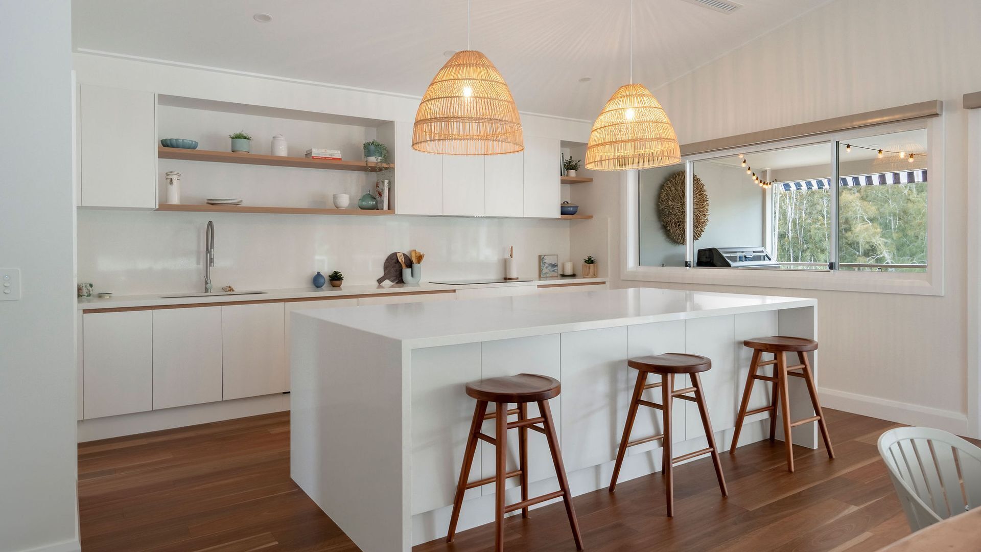 A modern kitchen with white cabinetry, a central island with stools, pendant lighting, and a view through a large window.