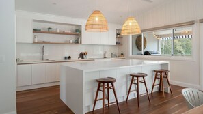 A modern kitchen with white cabinetry, a central island with stools, pendant lighting, and a view through a large window. - Holiday Mode | Lakefront Luxury with private jetty (Lake Conjola)