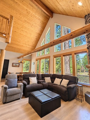 A spacious living area with a large sectional sofa, a coffee table, and a dining table. The room features high ceilings with exposed wooden beams, large windows offering views of trees, and a stone accent wall.