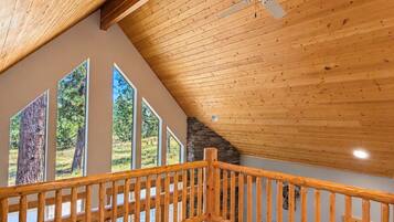 A spacious room with a sloped ceiling, a wooden ceiling fan, a view of trees through large windows, and a wooden railing along the balcony.
