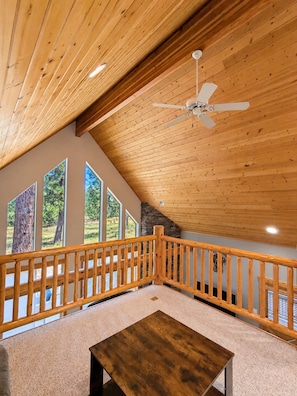 A spacious room with a sloped ceiling, a wooden ceiling fan, a view of trees through large windows, and a wooden railing along the balcony.