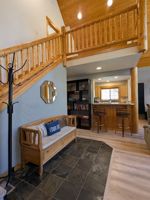 A wooden staircase leading to an upper level, a wooden bench with a cushion, a tiled floor, and a kitchen area with a bar.
