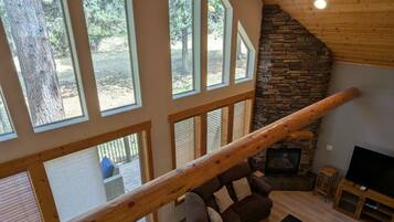 A spacious living room with a stone fireplace, large windows, and a wooden ceiling.