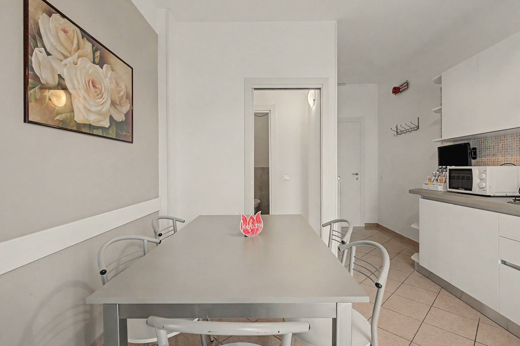 A modern kitchen with a microwave, a coffee machine, and a sink. There is a dining table with chairs and a red 'Welcome' sign.