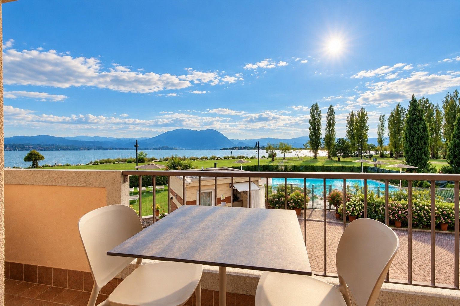 A balcony with a table and chairs, overlooking a pool and a landscaped garden with a mountain view.