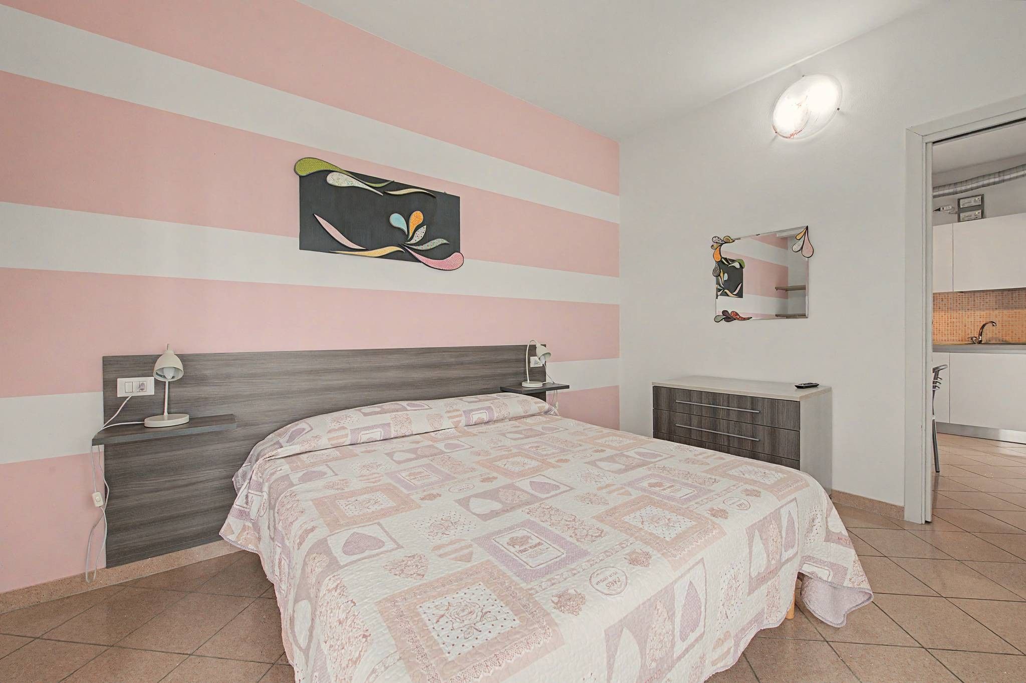 Ein Schlafzimmer mit einem Bett, Nachttischen, einem Schminktischchen, einer Kommode, einem Spiegel und einer Wand mit Streifenmuster.