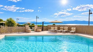 A swimming pool with lounge chairs and umbrellas, surrounded by a wooden fence, with a view of mountains and a body of water in the background.