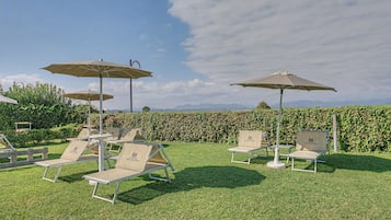 Ein Gartenbereich mit Sitzmöglichkeiten und Sonnenschirmen, umgeben von einer Hecke und Blick auf ferne Berge.