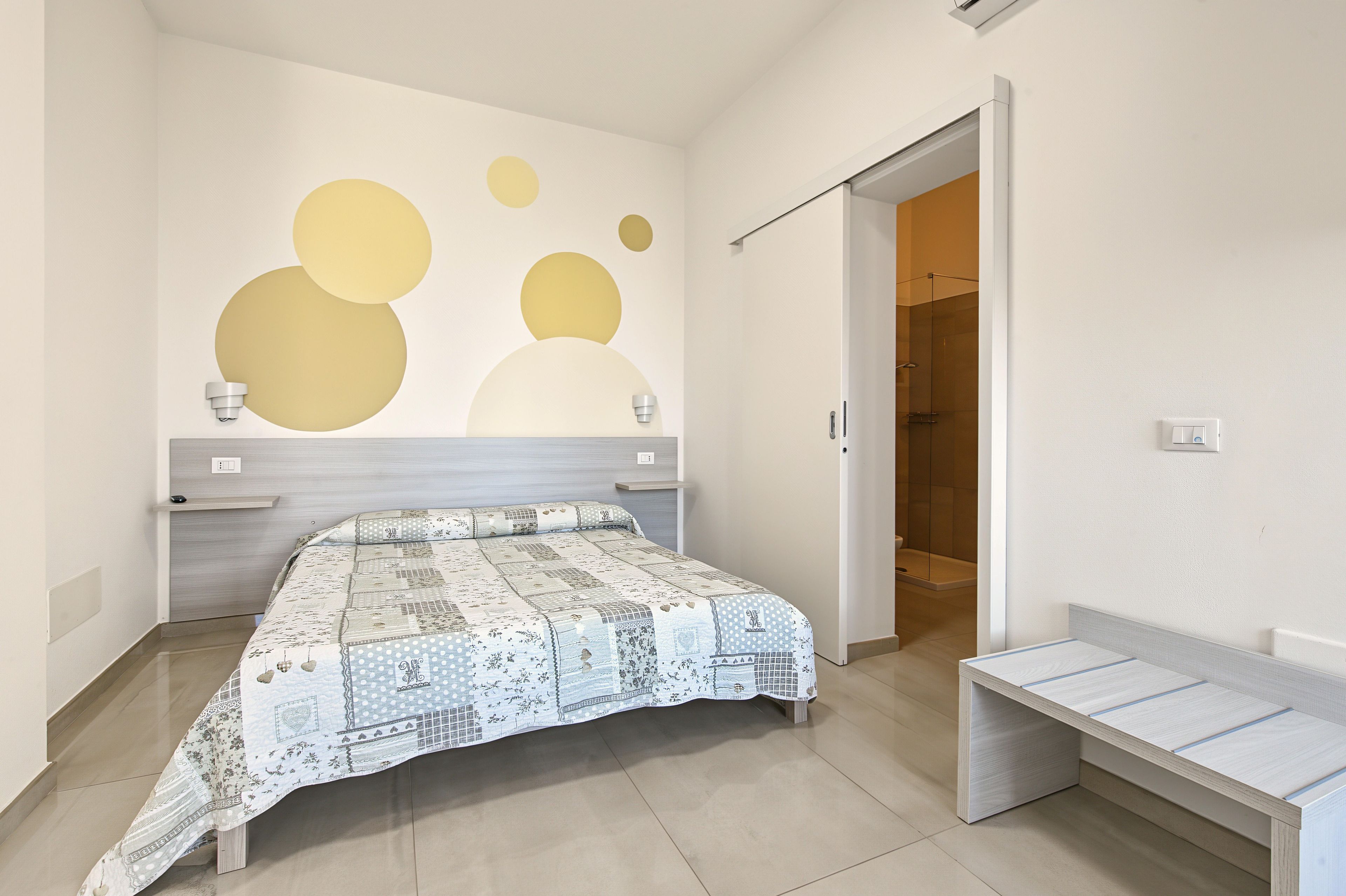 Un dormitorio con cama, una banca, una pared con arte abstracto y un baño visible a través de una puerta abierta.