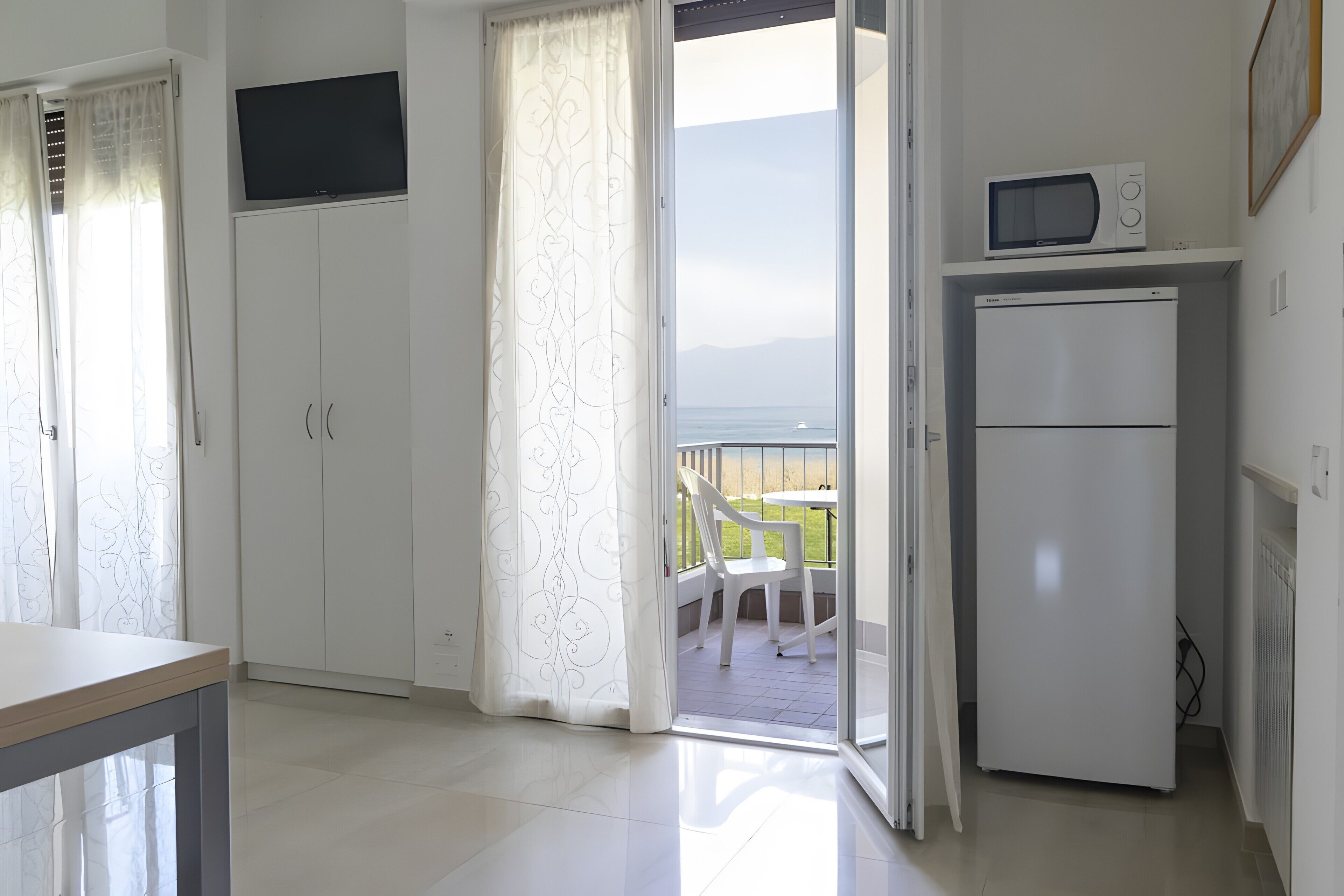 Ein Zimmer mit Balkon, einem weißen Kühlschrank, einer Mikrowelle und einem Fernseher an der Wand.