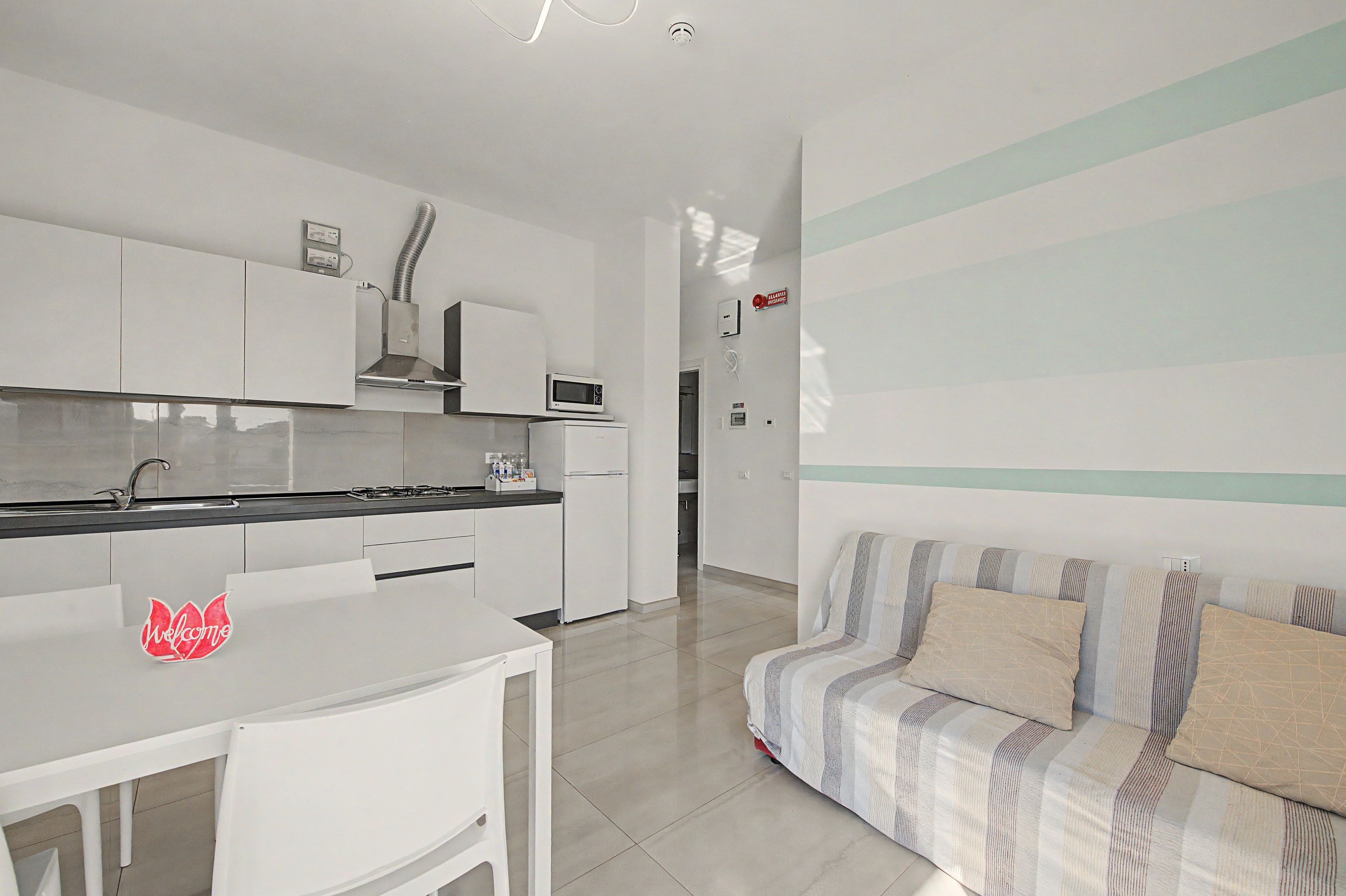 A modern kitchen with white cabinetry, a gas stove, and a microwave. There is a dining table with a 'Welcome' sign. A striped sofa is in the living area with a view of the kitchen.