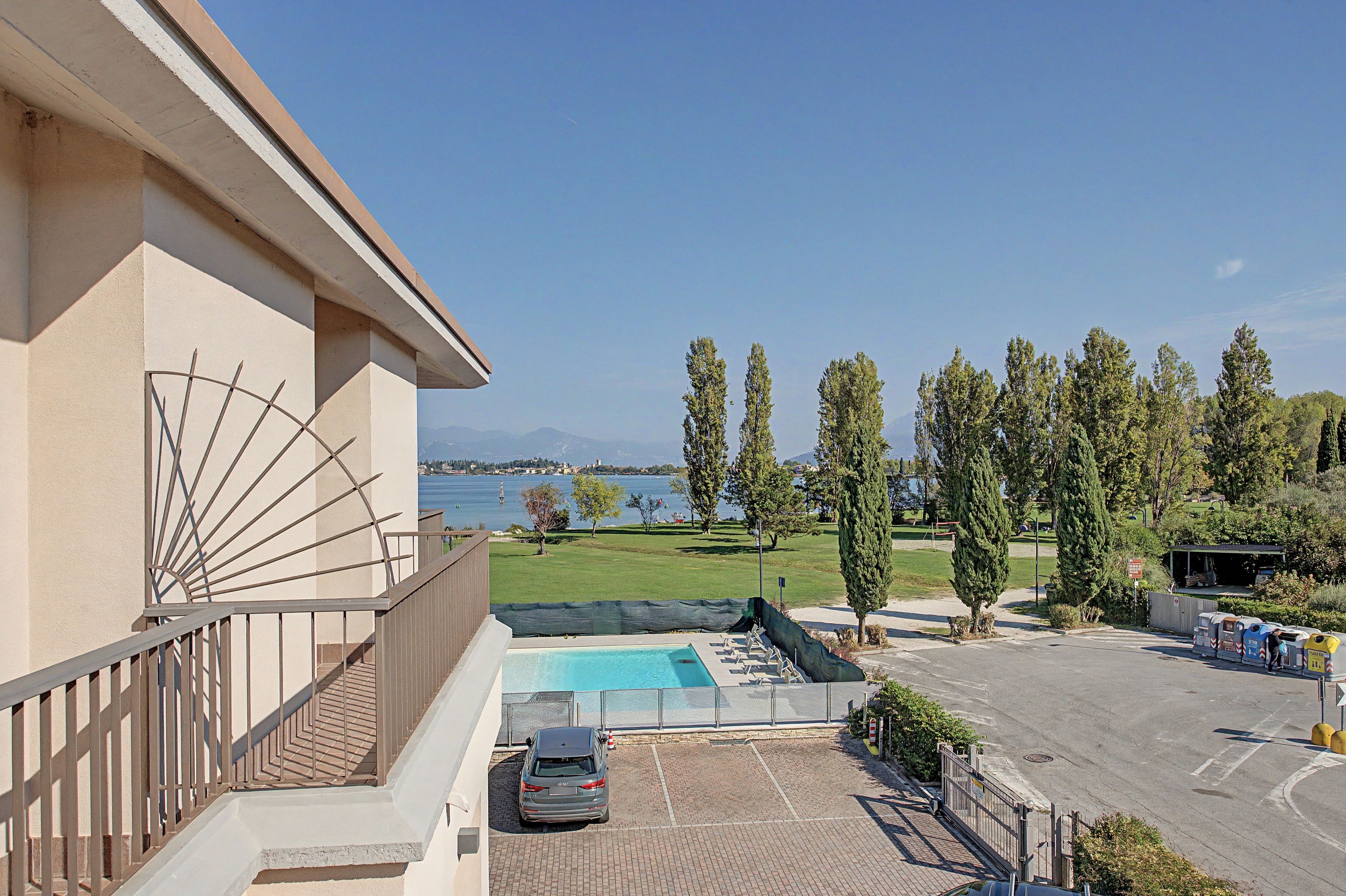 A pool area with a car parked nearby, a view of the sea, and a building with a balcony.
