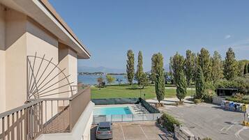 A pool area with a car parked nearby, a view of the sea, and a building with a balcony.