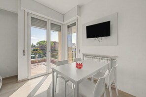 A modern room with a white table, chairs, a TV, and a balcony with a view of buildings. - Papavero Apartment - Lake View Three-Room with Wi-Fi and Air Conditioning (Sirmione)