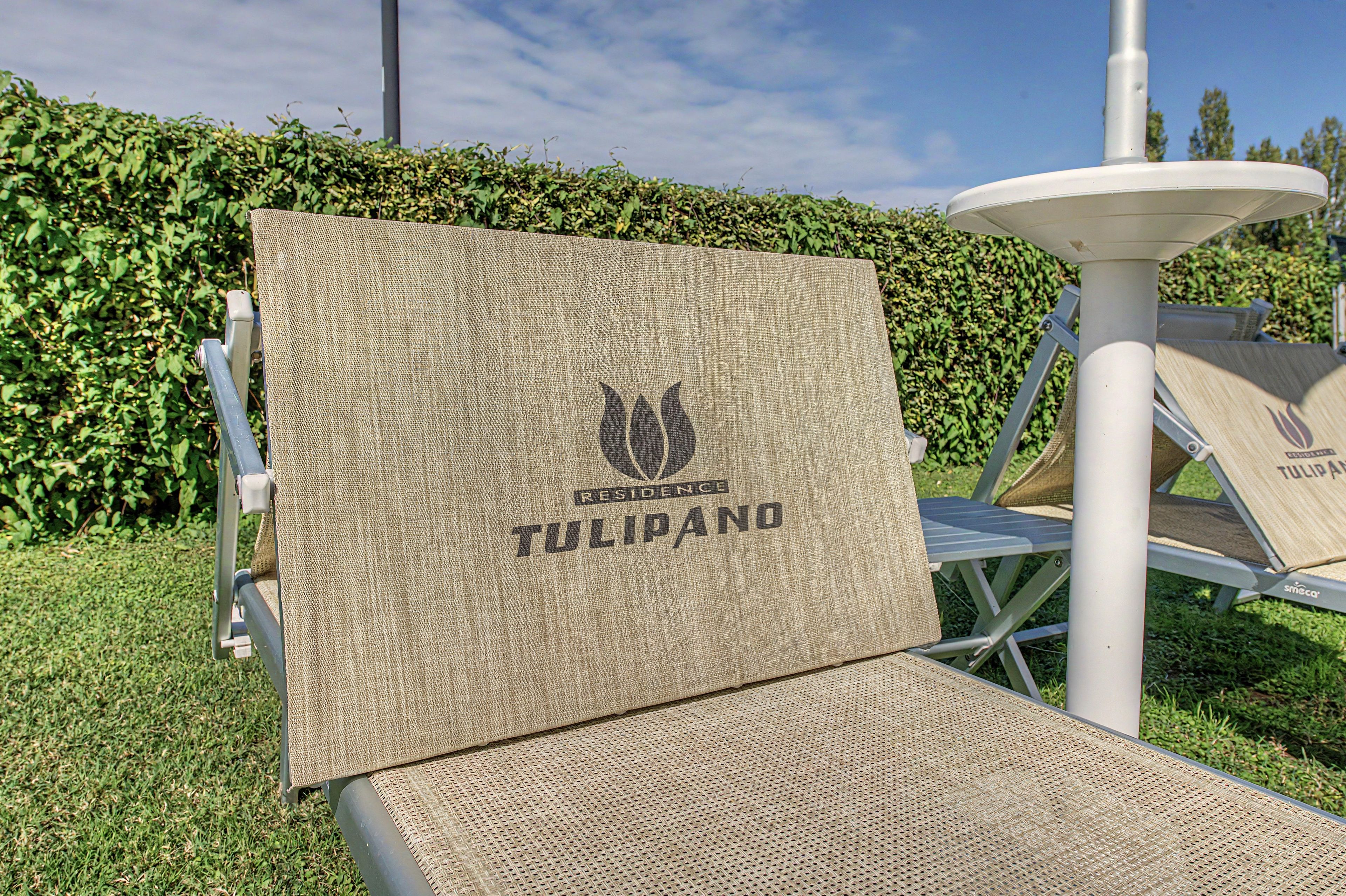 Ein Stuhl mit gewobener Sitzfläche und Rückenlehne, gekennzeichnet mit dem Logo 'RESIDENCE TULIPANO', befindet sich draußen vor einer Hecke und einem weißen Gebäude.