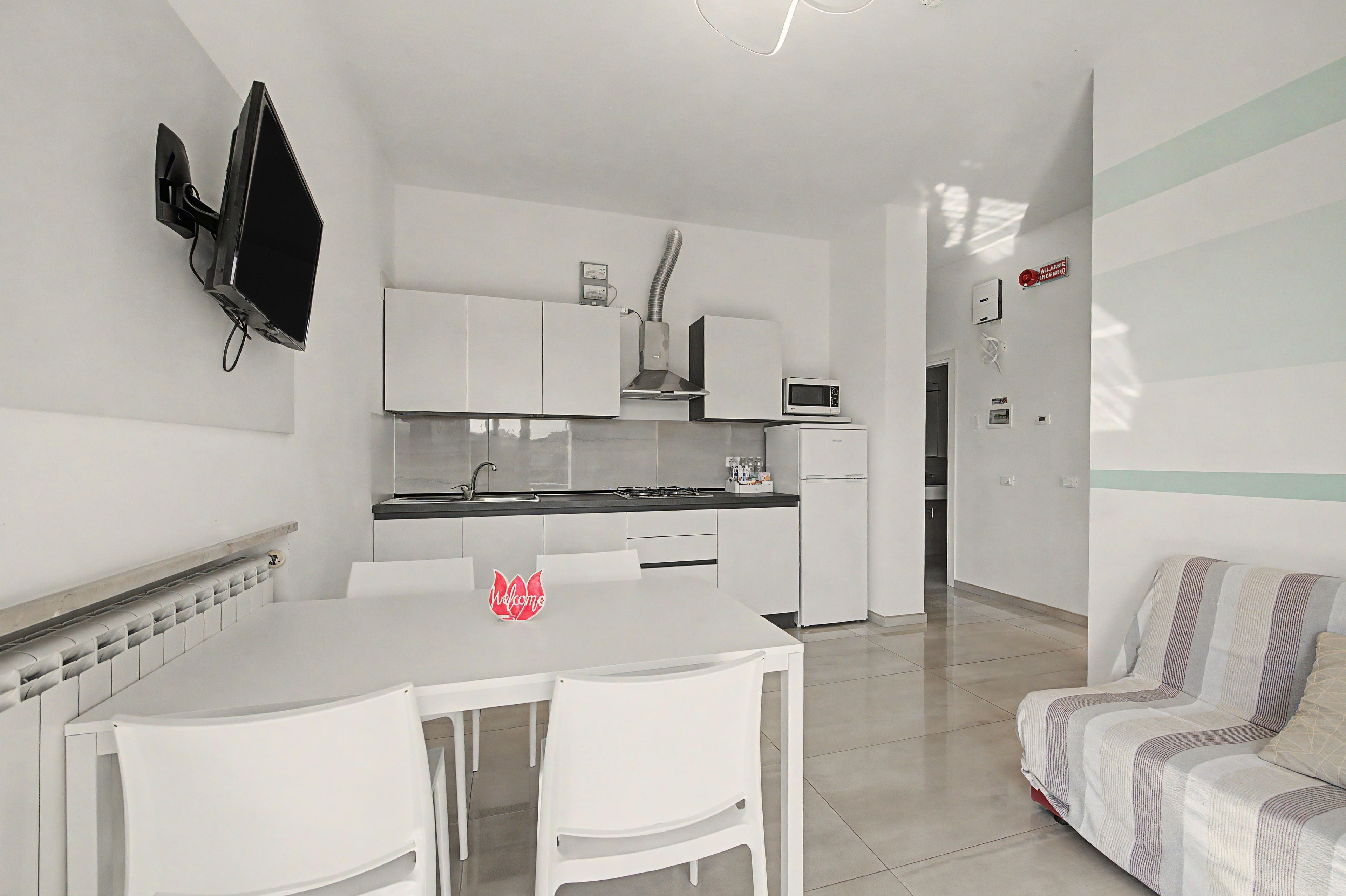 A modern kitchen with white cabinetry, a microwave, and a gas stove. There is a white dining table with chairs and a red 'Welcome' sign. A sofa is visible in the adjacent living area.