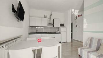 A modern kitchen with white cabinetry, a microwave, and a gas stove. There is a white dining table with chairs and a red 'Welcome' sign. A sofa is visible in the adjacent living area.
