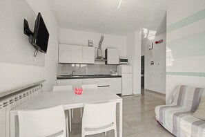 A modern kitchen with white cabinetry, a microwave, and a gas stove. There is a white dining table with chairs and a red 'Welcome' sign. A sofa is visible in the adjacent living area. - Papavero Apartment - Lake View Three-Room with Wi-Fi and Air Conditioning (Sirmione)