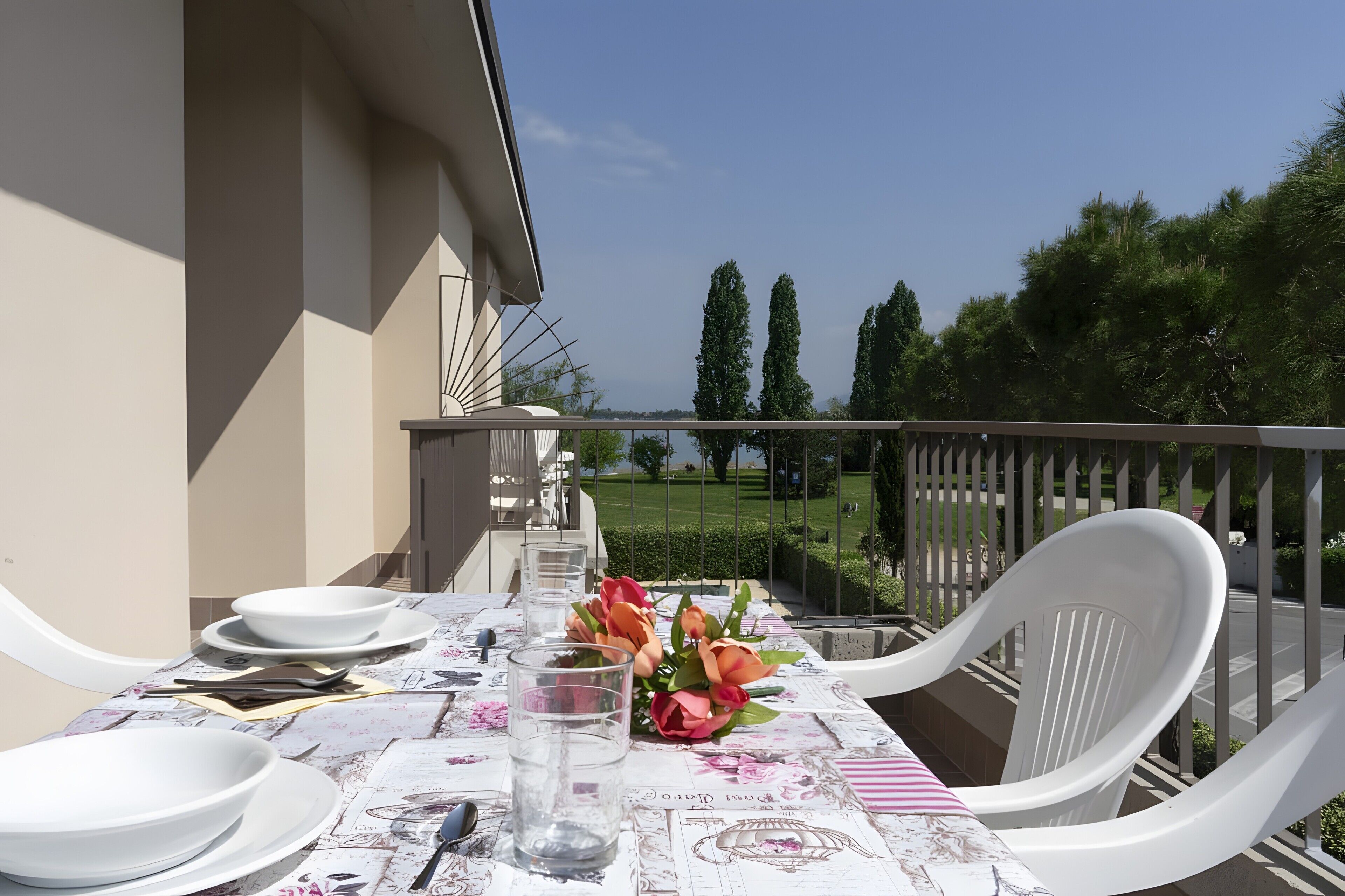 Une table de balcon, dressée avec des assiettes, des couverts et un verre d’eau, donnant sur un parc arboré et un ciel dégagé.