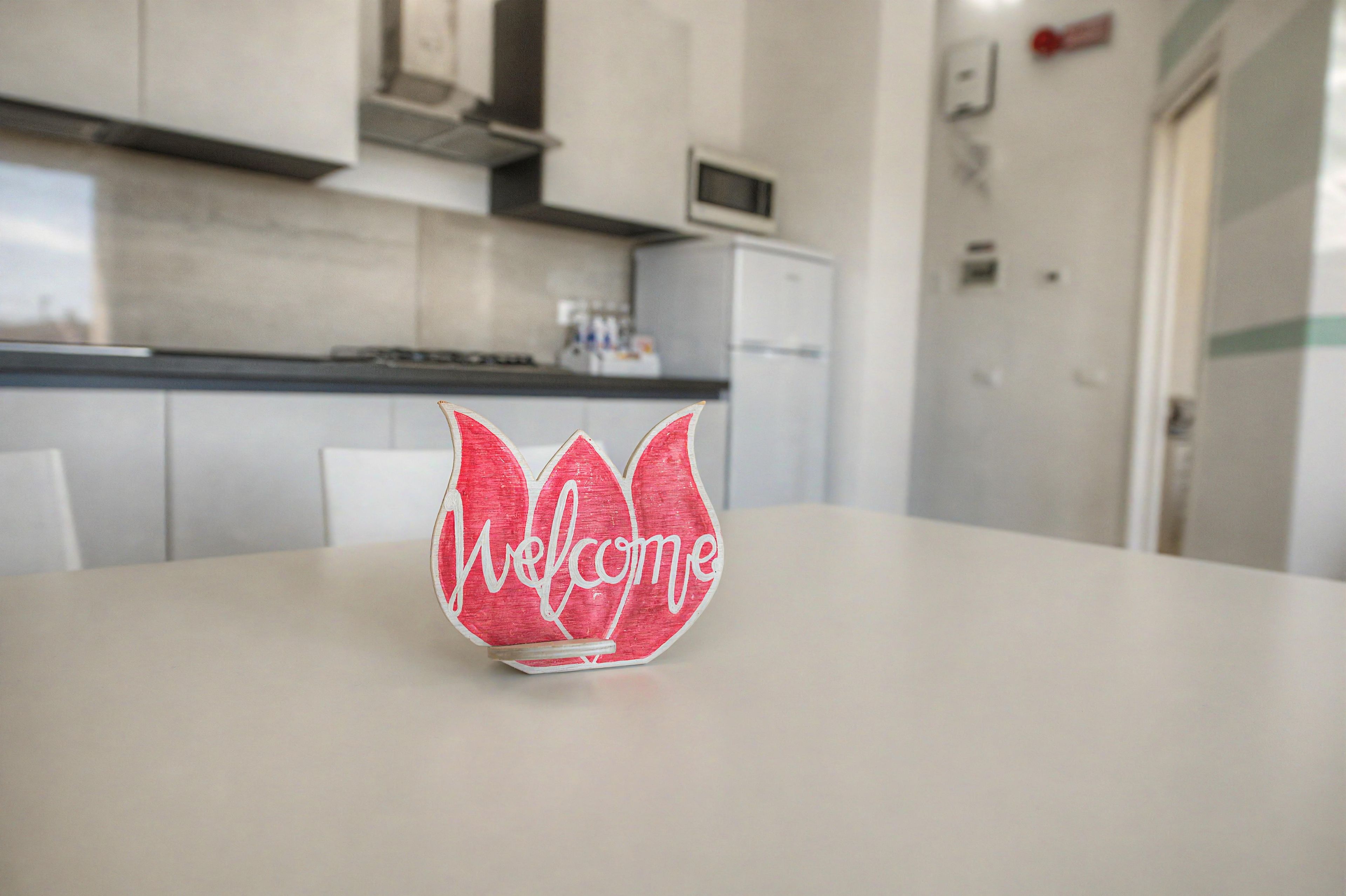 A red and white paper welcome sign on a table in a modern kitchen with white cabinets and a microwave.