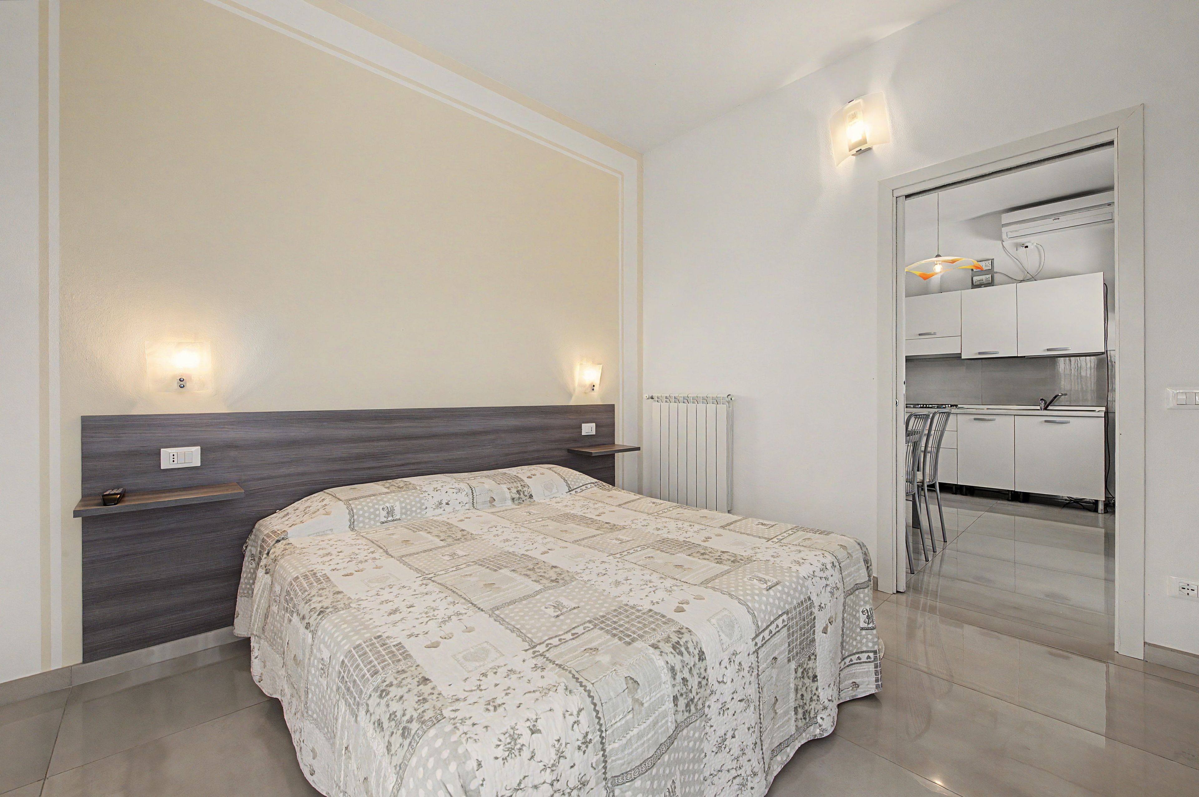 Ein Schlafzimmer mit Bett, Nachttischen, einem Heizkörper und Blick in einen Küchenbereich.