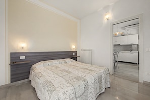 Ein Schlafzimmer mit Bett, Nachttischen, einem Heizkörper und Blick in einen Küchenbereich.