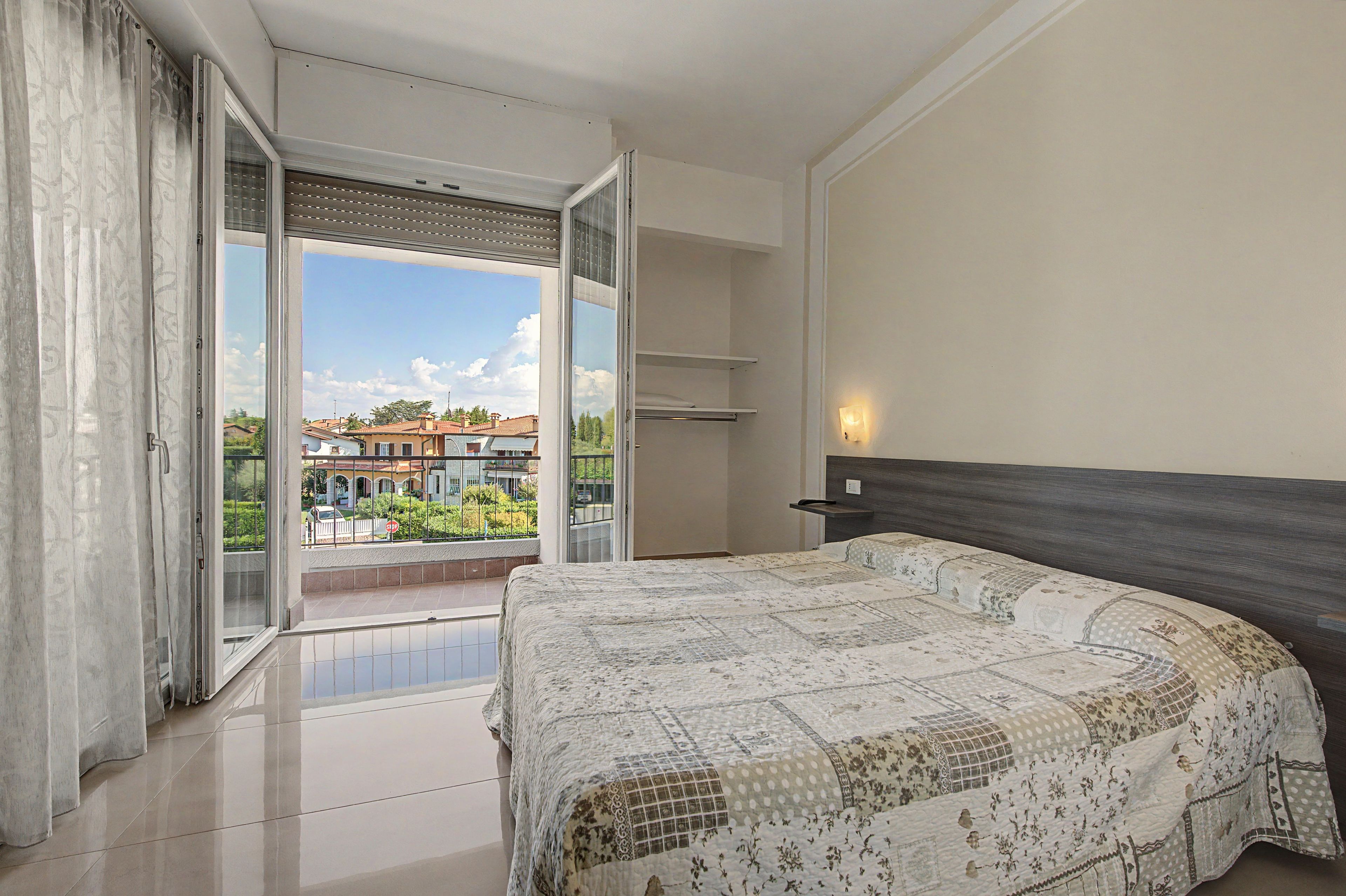 A bedroom with a large bed, a sliding glass door to a balcony, and a view of a residential area.