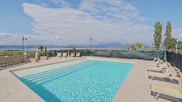 Una piscina rettangolare con acqua cristallina, circondata da un solarium piastrellato. Sono presenti lettini e un'area relax coperta con ombrellone. La piscina si staglia sullo sfondo di un cielo sereno e montagne in lontananza.