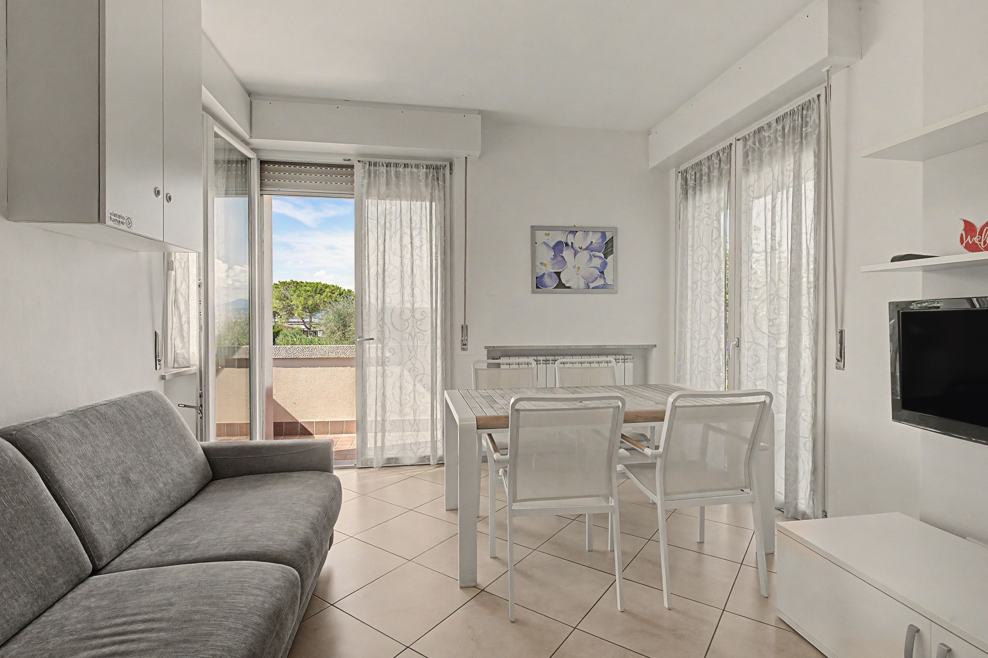 Ein modernes Wohnzimmer mit einem grauen Sofa, einem Esstisch mit Stühlen, einem Fernseher und einem Balkon mit Blick auf Grünflächen.