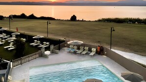 Una piscina con lettini e ombrelloni, circondata da un prato e con vista sul tramonto sull'acqua.