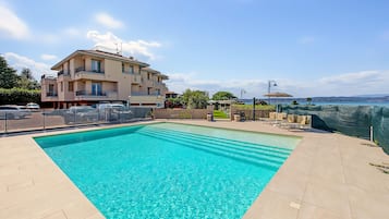 Una piscina con agua cristalina azul, rodeada por una solana con baldosas, un edificio con balcones y vistas al mar.