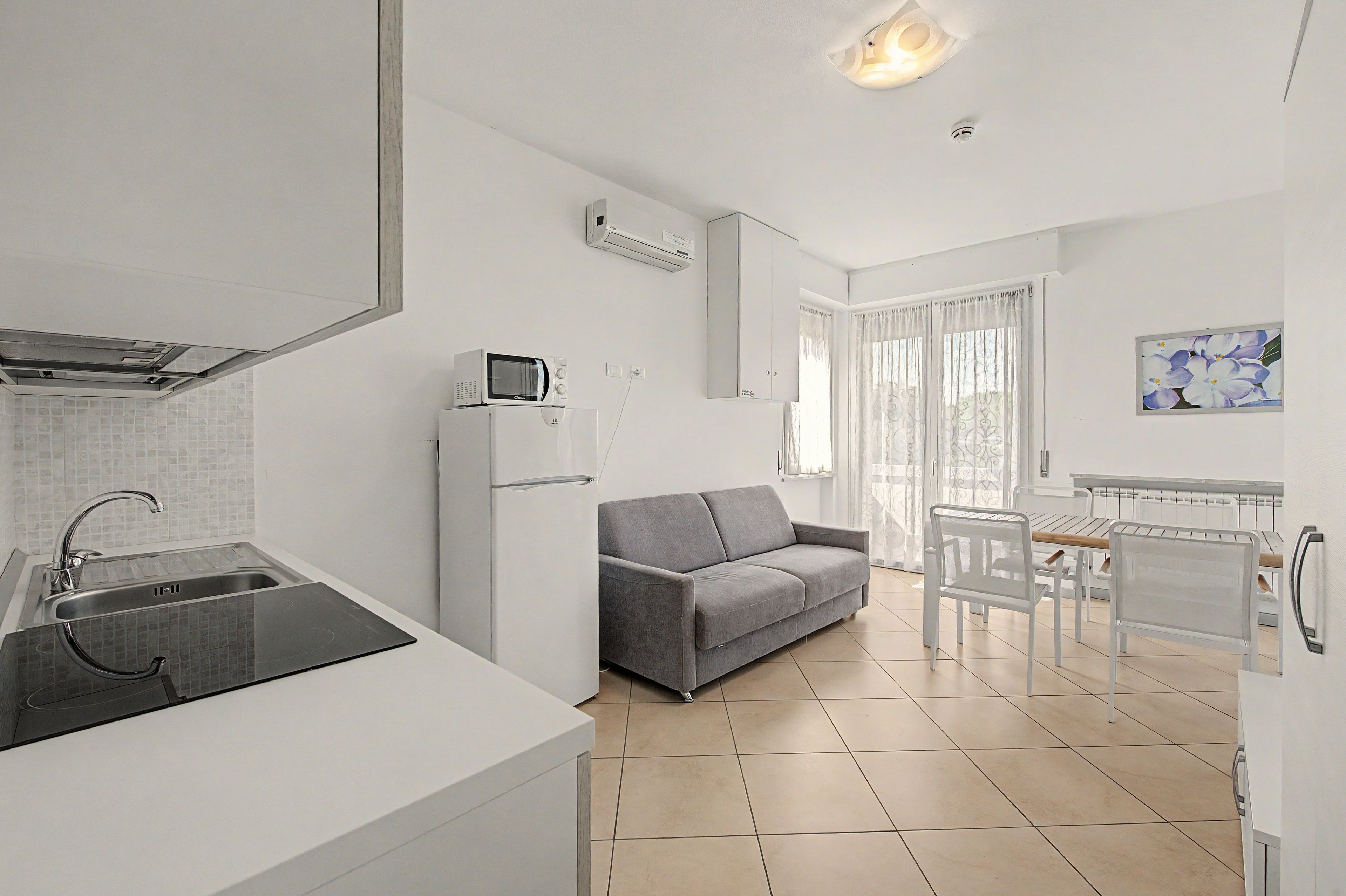 A compact living space with a kitchenette, a gray sofa, a dining table with chairs, and a painting on the wall.