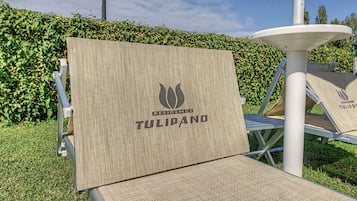 Una silla con asiento y respaldo trenzados, con el logotipo 'RESIDENCE TULIPANO', ubicada al aire libre con un seto y una estructura blanca al fondo.