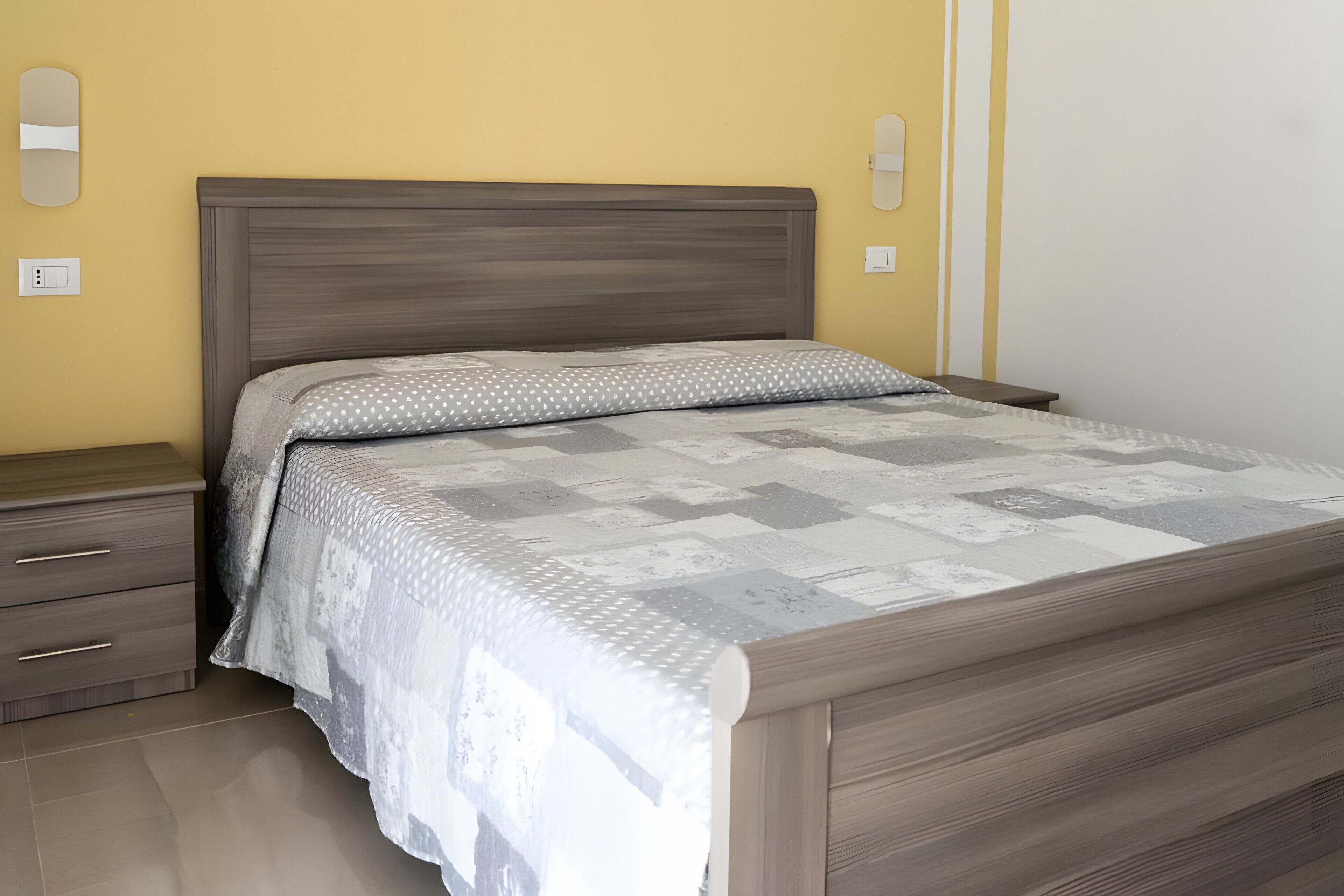 A bedroom with a bed, a nightstand, a drawer, and a wall-mounted light switch.