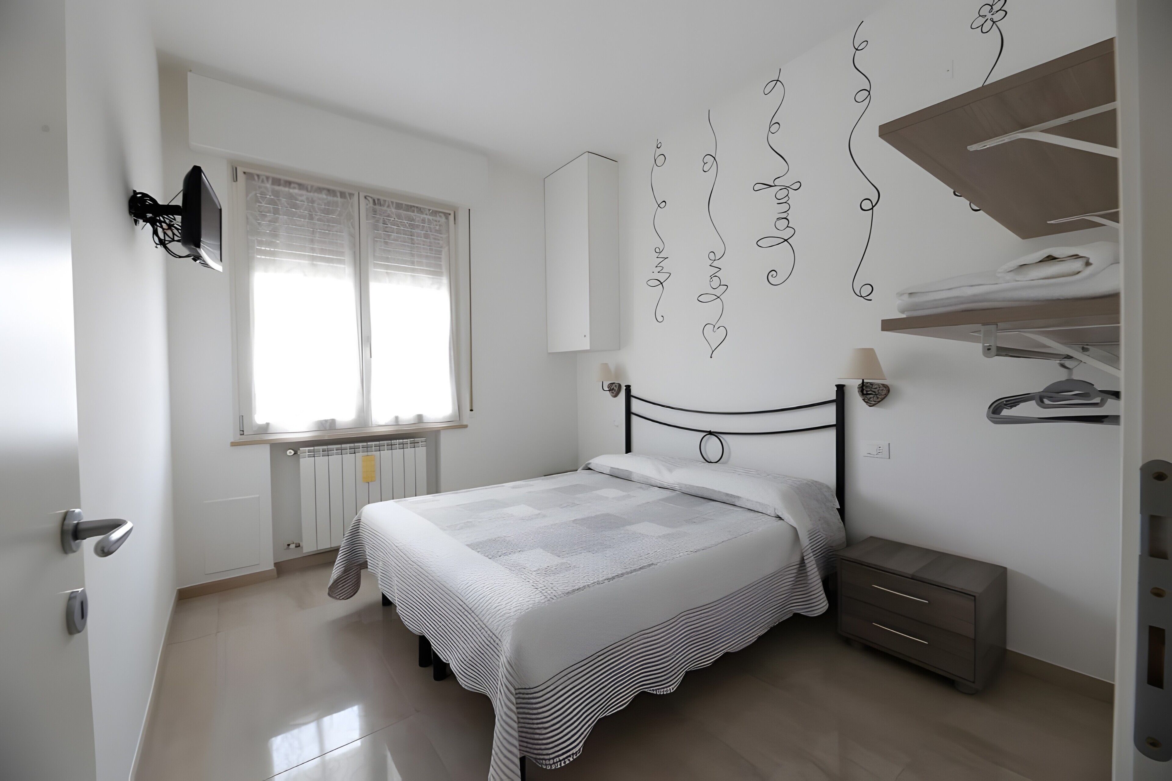 Un dormitorio con cama, radiador, ventana con persianas, estante con ropa doblada y mesita de noche con lámpara.