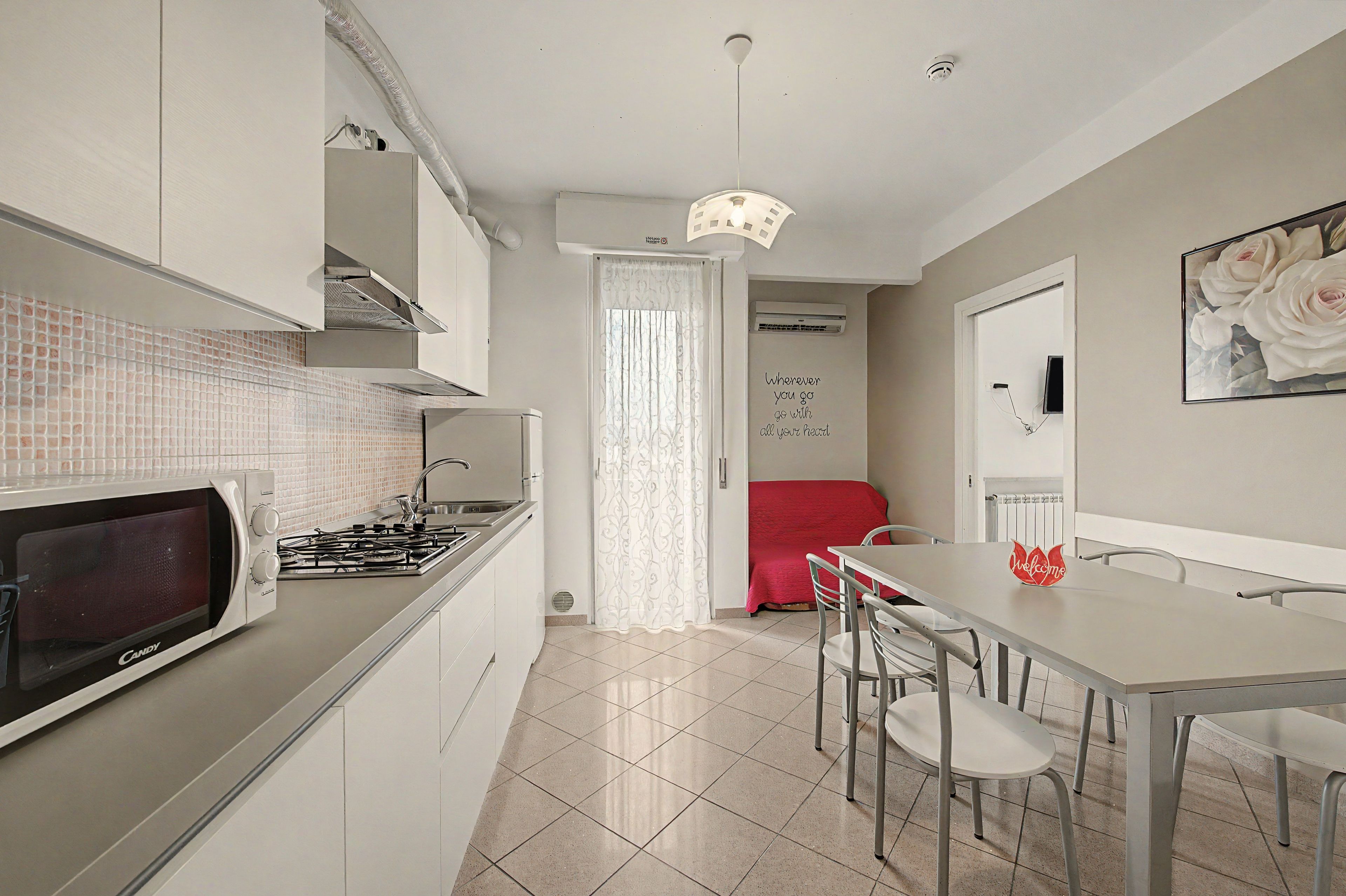 Une cuisine compacte équipée d’un four à micro-ondes, d’une cuisinière et de meubles blancs. On y trouve un coin repas avec une table et des chaises, et une chaise rouge à l’arrière-plan.