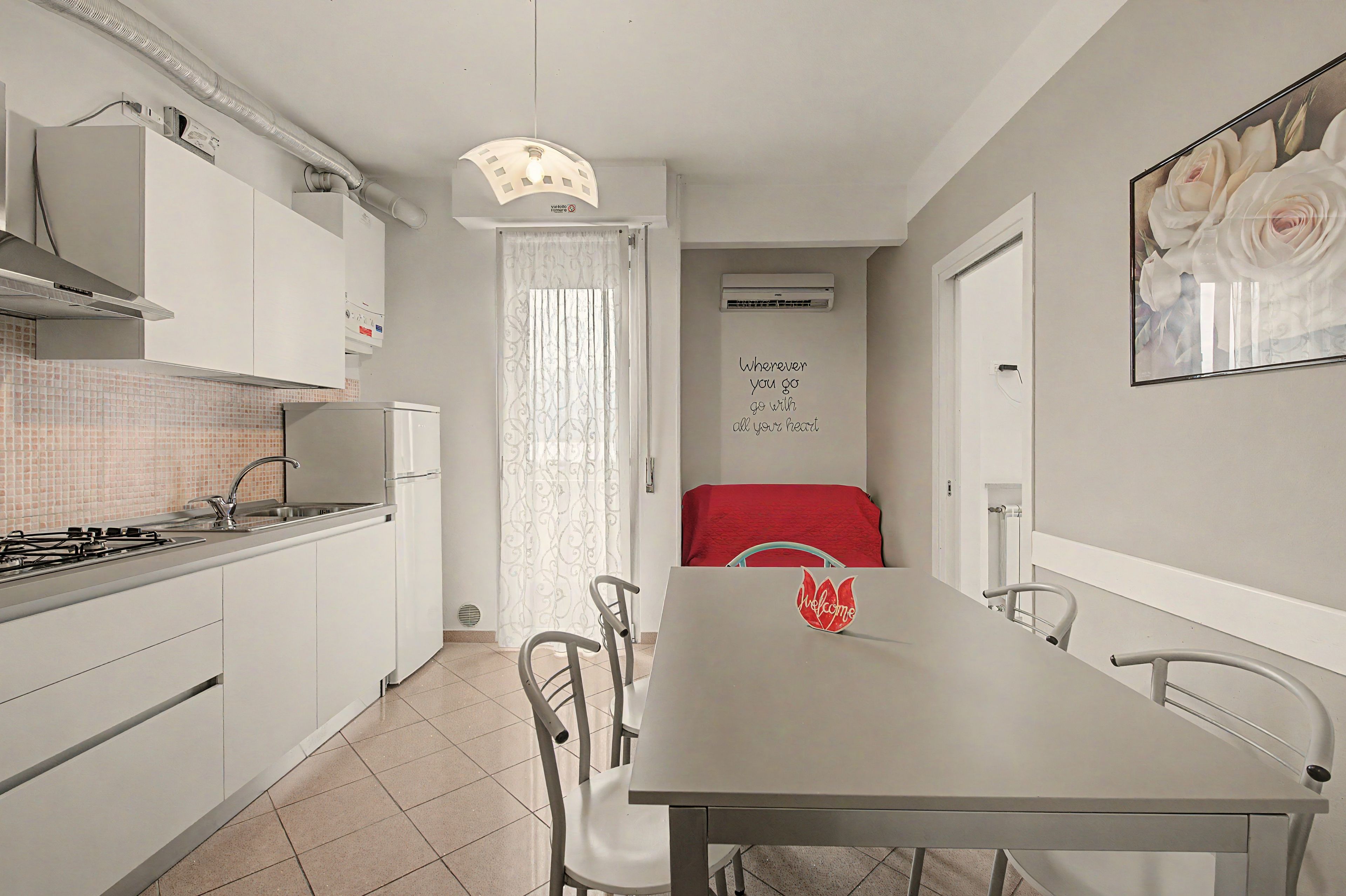 Cocina compacta con estufa a gas, lavamanos y armarios blancos. Una mesa de comedor con sillas está ubicada en el centro, y se ve una cama roja al fondo.