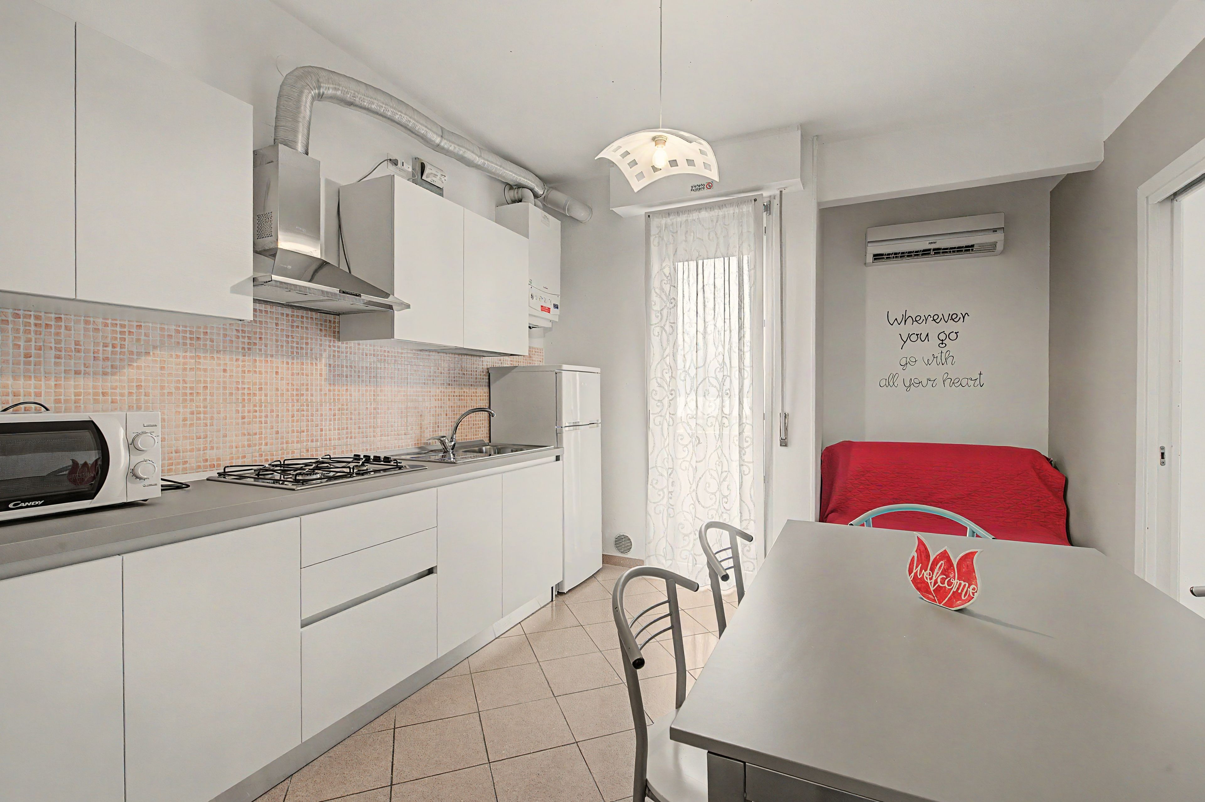 Cocina compacta con armarios blancos, estufa a gas, microondas y una mesa roja con un cartel de 'Welcome'.