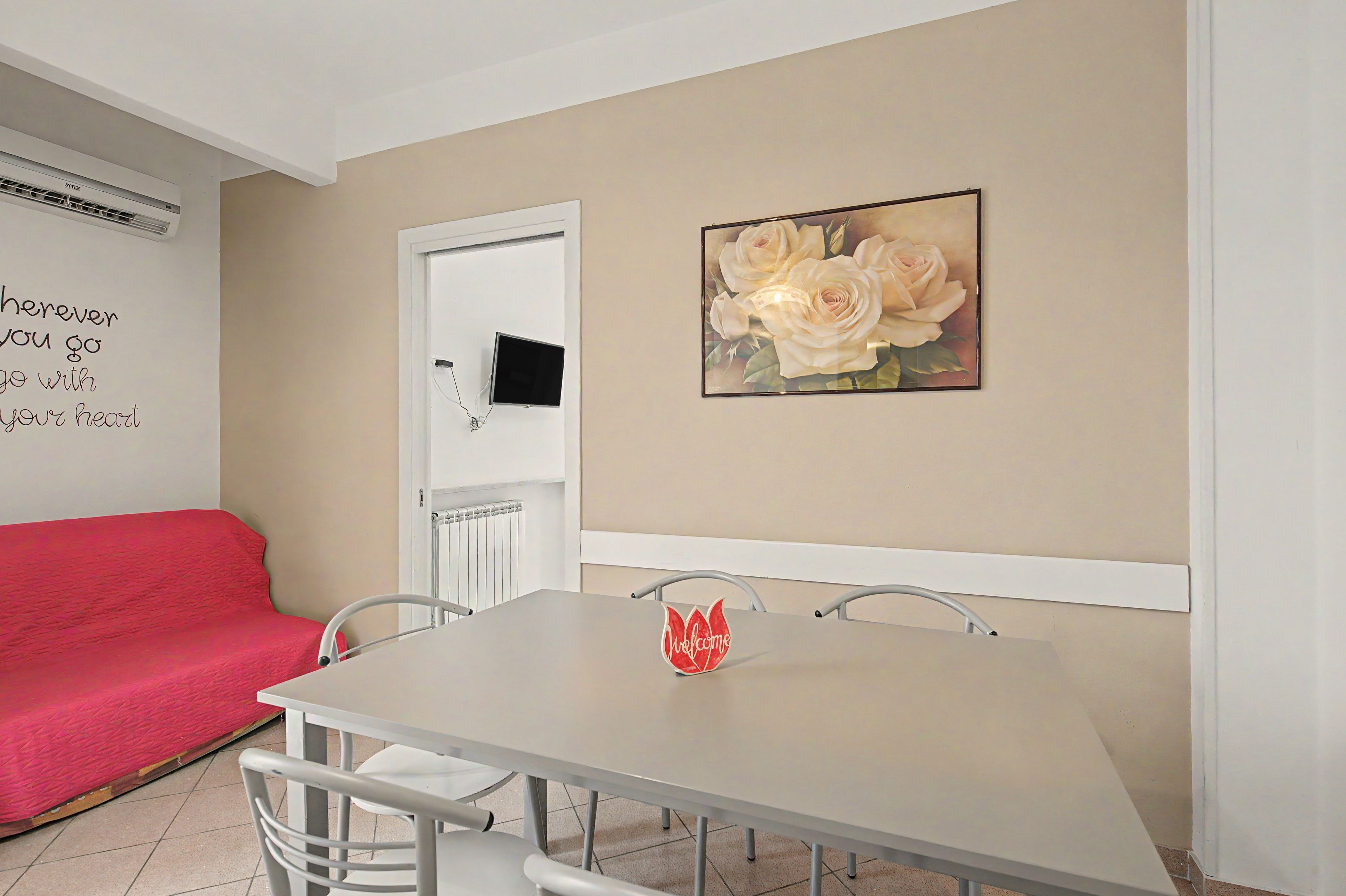 Ein Essbereich mit Tisch, Stühlen, einem roten Sofa, einem an der Wand befestigten Fernseher und einem floralen Bild in einem Rahmen.
