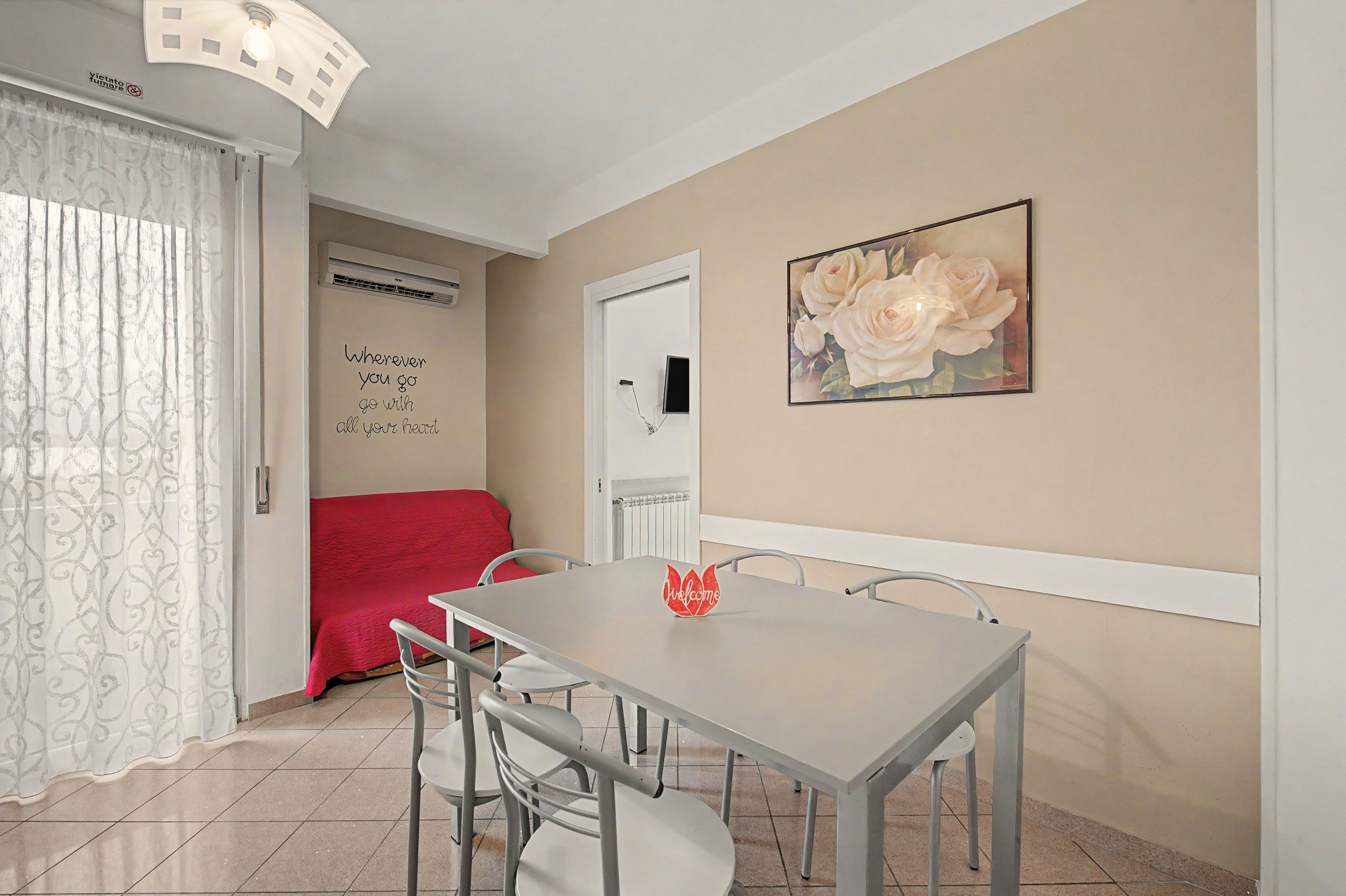 A dining area with a table and chairs, a red sofa, a wall with a rose painting, and a door with a sign.