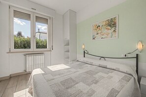 Ein Schlafzimmer mit einem Bett, einem Heizkörper, einem Fenster mit Jalousien und einem an der Wand befestigten Regal.