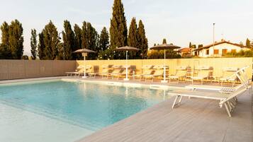Una piscina con sillas de descanso y sombrillas, rodeada por una terraza de madera y árboles altos.