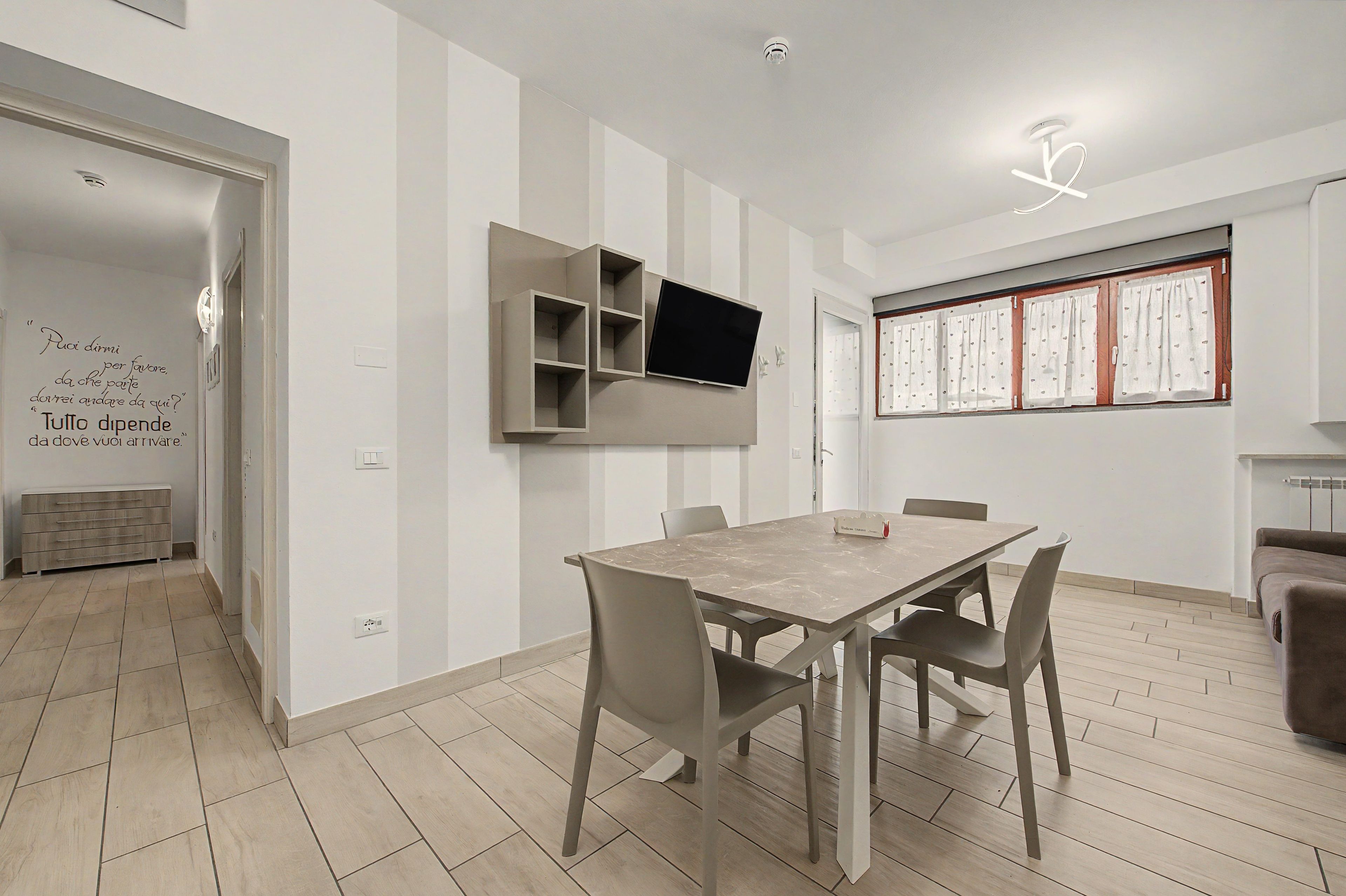 Un comedor moderno con una mesa rectangular y cuatro sillas, un televisor de pantalla plana montado en la pared, y un ventanal con cortinas.
