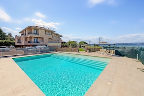 A swimming pool with a clear blue water, surrounded by a tiled deck, a building with balconies, and a view of the sea. - Ninfea Apartment – Superior Four-Room with Shared Pool, WiFi, and Air Conditioning (Sirmione)