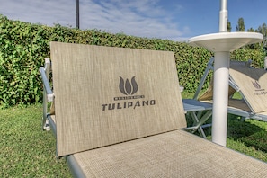 A chair with a woven seat and backrest, branded with 'RESIDENCE TULIPANO' logo, positioned outdoors with a hedge and a white structure in the background. - Ninfea Apartment – Superior Four-Room with Shared Pool, WiFi, and Air Conditioning (Sirmione)