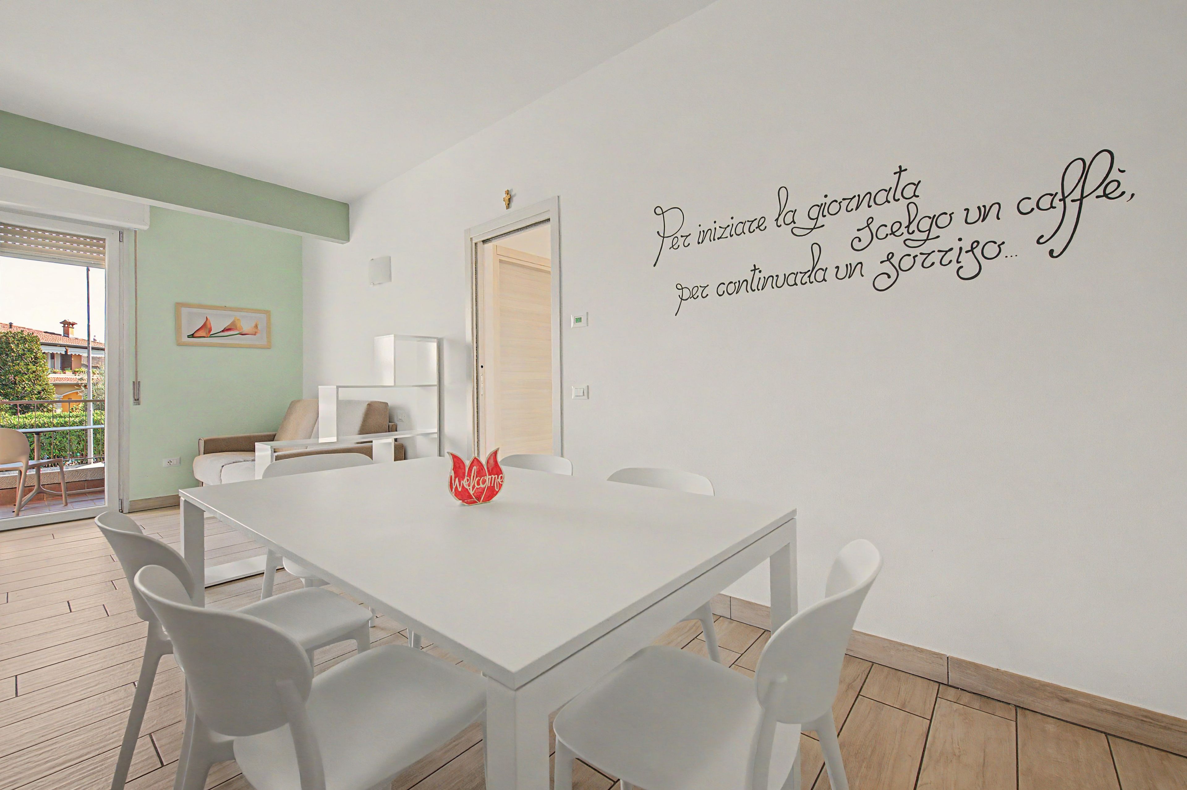 A dining area with a white table and chairs, a red 'Welcome' sign, and a wall with a motivational quote in Italian.